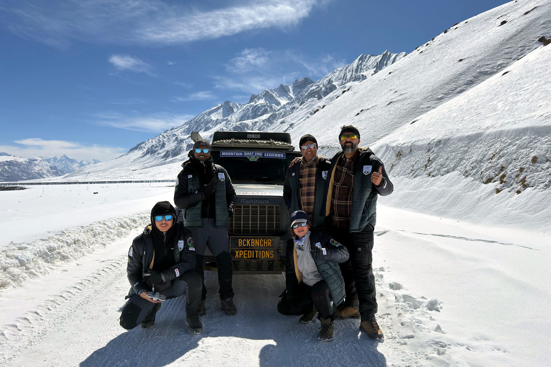 The QDE team beside their expedition vehicle on a snowbound Spiti road, snow peaks behind